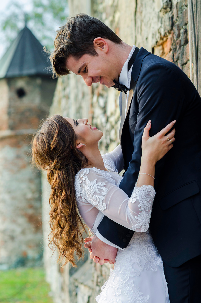 Uśmiechnięta para młoda w objęciach na tle kamiennej ściany z wieżą w oddali, panna młoda w koronkowej sukni z długimi rękawami, pan młody w czarnym smokingu z muszką.