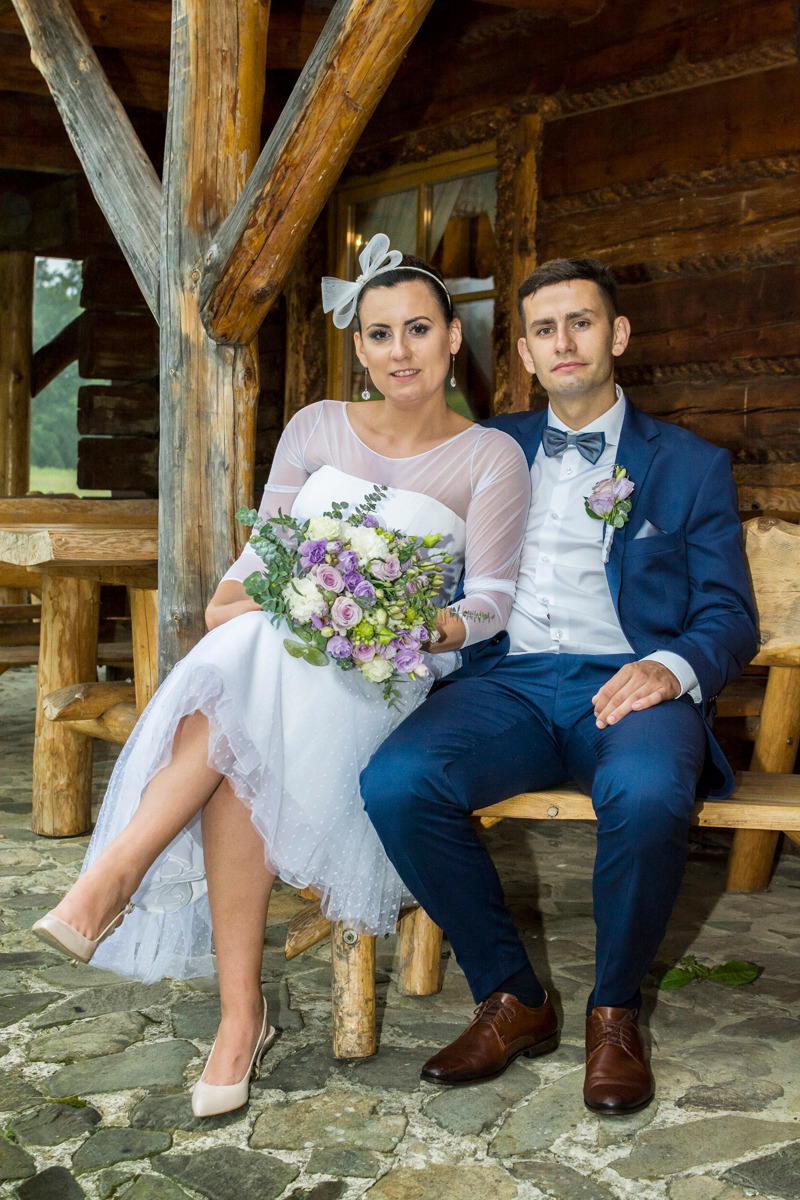 Para młoda siedząca na drewnianej ławce przed drewnianą konstrukcją. Panna młoda w białej sukni z bukietem fioletowych i białych kwiatów, pan młody w granatowym garniturze.