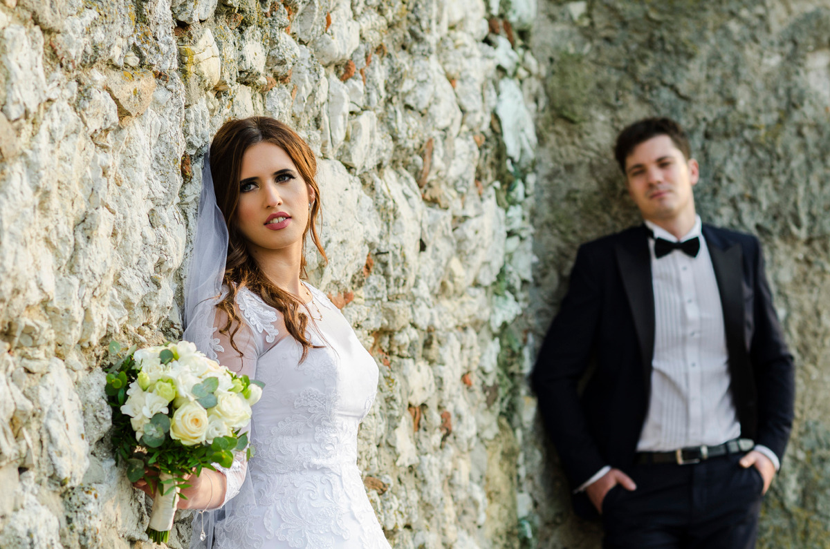 Panna młoda w koronkowej sukni ślubnej z bukietem białych róż oparta o kamienną ścianę, obok pan młody w czarnym smokingu z muchą.