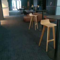 Ciemna wykładzina w dużej przestrzeni biurowej z drewnianymi stołkami barowymi i miękkimi siedziskami tworzy nowoczesny, minimalistyczny design.