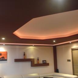 GipsoFarb-Robert Kuściński - Sufit podwieszany z oświetleniem LED i brązowymi elementami dekoracyjnymi w nowoczesnym salonie. Widoczne półki z dekoracjami i obraz na ścianie.