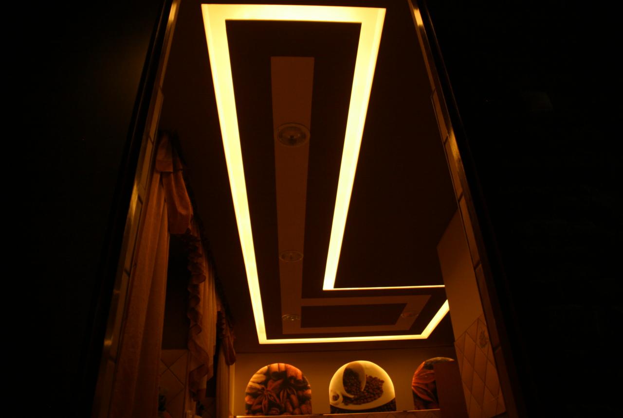 Nowoczesny sufit podwieszany z oświetleniem LED w kształcie litery 'Z', widziany z dołu. Dekoracyjne półki z motywami kawowymi w tle. Ciemne otoczenie.