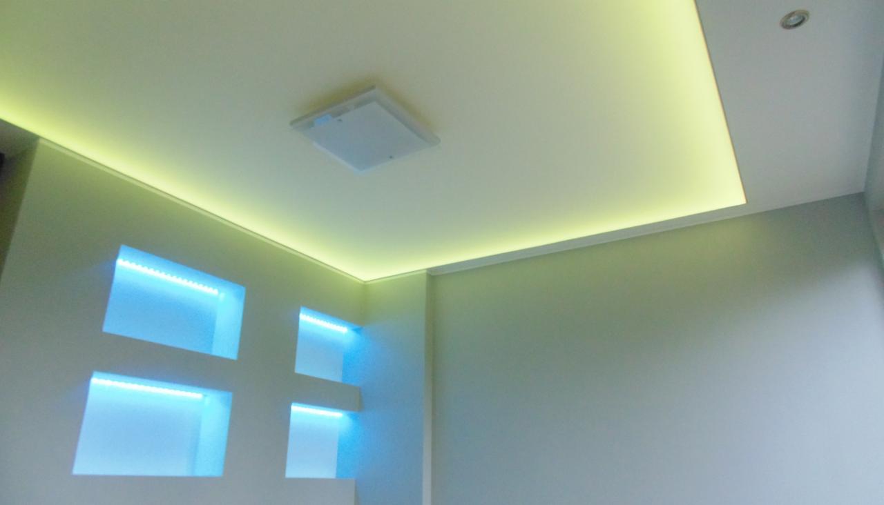 Nowoczesny sufit podwieszany z oświetleniem LED: żółte obwodowe i niebieskie wnękowe, tworzące krzyż. Białe ściany i centralny właz rewizyjny.