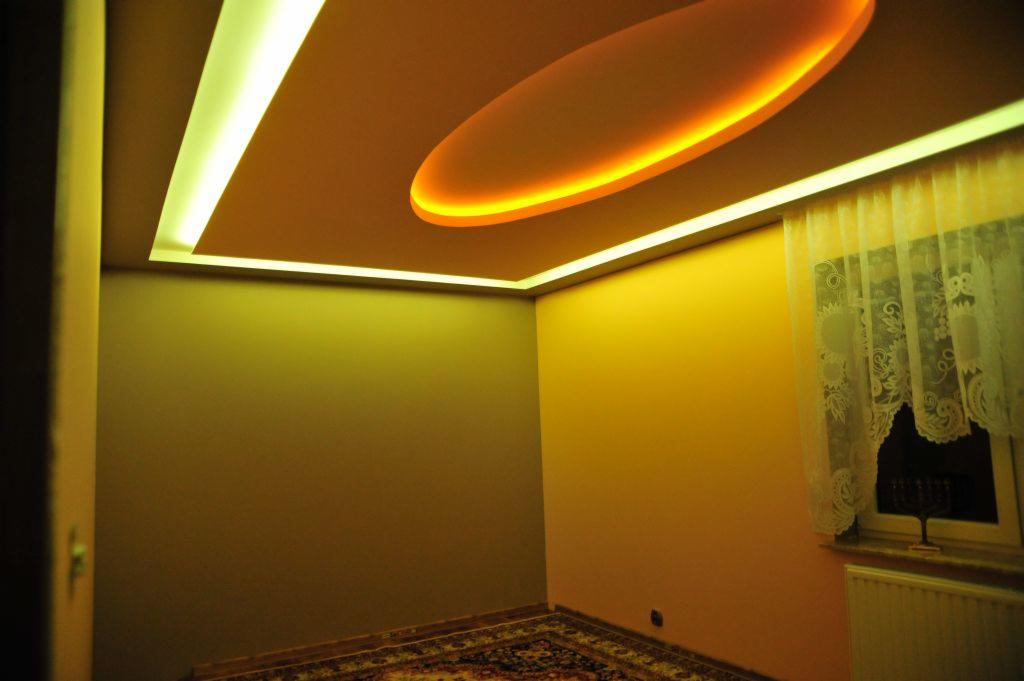 Pokój z podwieszanym sufitem z oświetleniem LED. Sufit w kolorze ciepłego beżu, ściany w odcieniach żółci i oliwki. Dywan z wzorem orientalnym.
