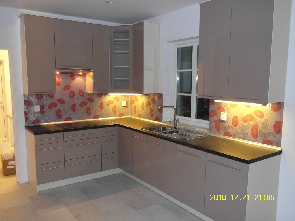 Jasna kuchnia z błyszczącymi, beżowymi szafkami, czarnym blatem i tapetą w czerwone maki. Widoczny zlew, płyta grzewcza i oświetlenie podszafkowe.