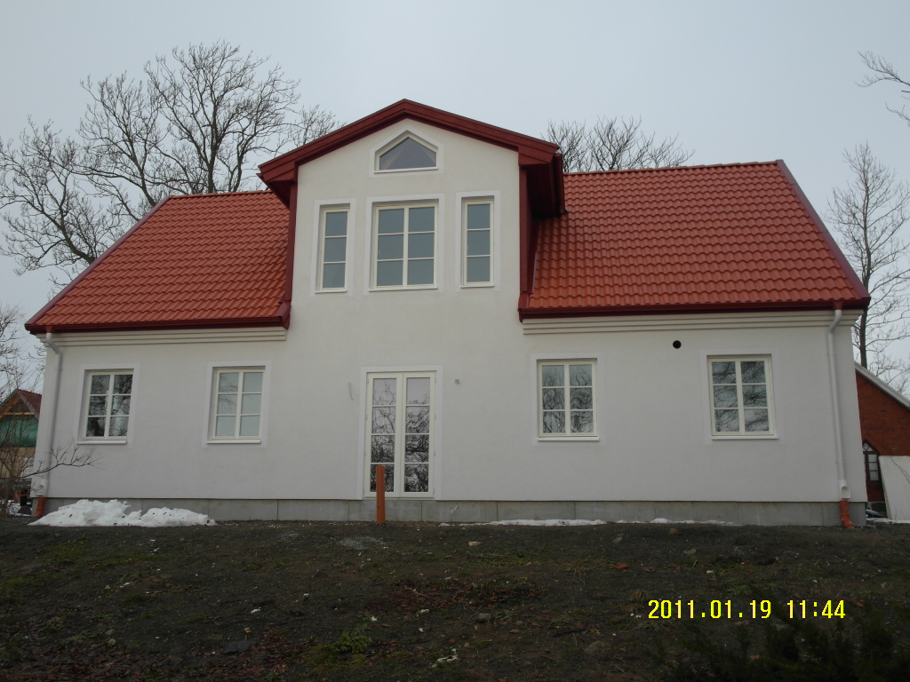 Dwukondygnacyjny dom z białymi ścianami i czerwoną dachówką, sfotografowany w pochmurny dzień. W oknach odbijają się gałęzie drzew bez liści. Na ziemi resztki śniegu.
