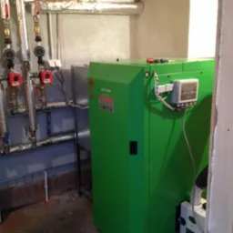 Zielony piec na paliwo stałe marki Kostrzewa w kotłowni, z widoczną instalacją hydrauliczną i elementami sterowania, oświetlony naturalnym światłem.