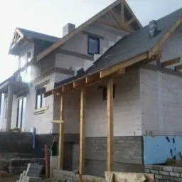 Budowa domu w stanie surowym otwartym z widoczną więźbą dachową, ściany z białego betonu komórkowego, częściowo pokryty dach, widoczne fundamenty i izolacja termiczna.