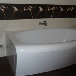 Biała wanna narożna z chromowaną baterią, obok grzejnik drabinkowy i ciemne płytki podłogowe, ściana wyłożona jasnymi płytkami z ciemnym pasem dekoracyjnym z motywem kwiatowym.