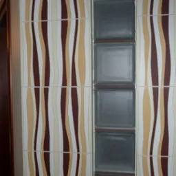 Ściana wyłożona płytkami z falistym wzorem w odcieniach brązu, beżu i bieli, sąsiadująca z pionowym pasem z luksferów w łazience.