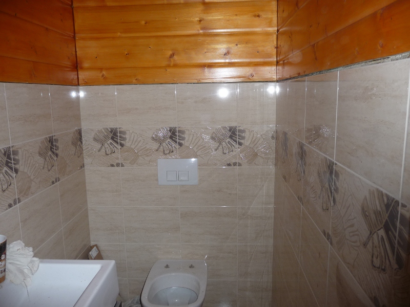 Wykończona łazienka z beżowymi płytkami z motywem liści, drewnianą boazerią sufitową i zainstalowaną białą toaletą oraz spłuczką podtynkową.