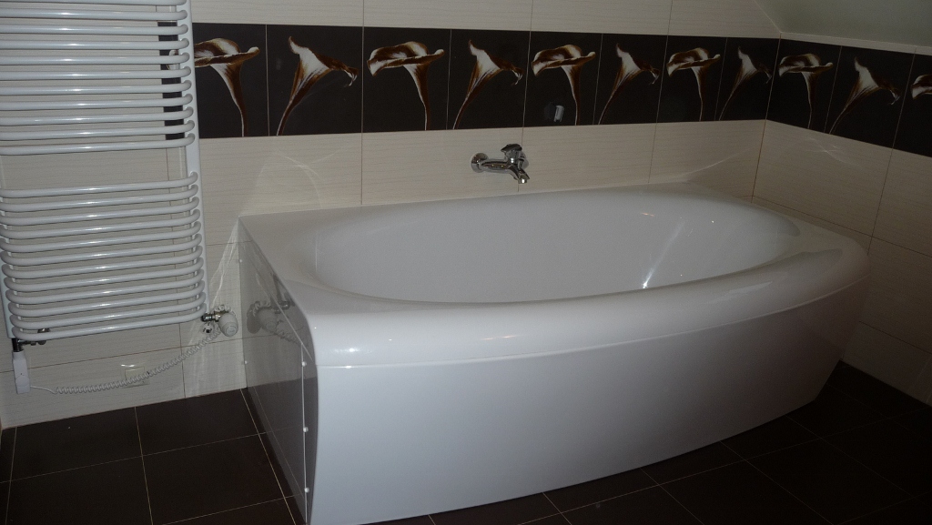 Biała wanna narożna z chromowaną baterią, obok grzejnik drabinkowy i ciemne płytki podłogowe, ściana wyłożona jasnymi płytkami z ciemnym pasem dekoracyjnym z motywem kwiatowym.