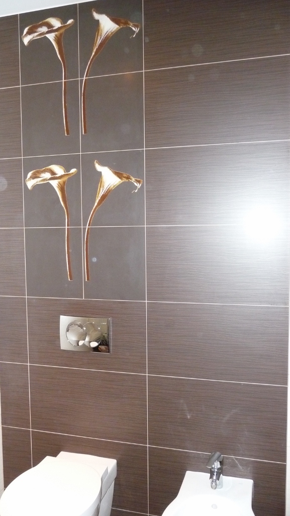 Brązowe płytki ceramiczne z motywem kwiatów kalii zdobią ścianę nad toaletą i bidetem w łazience, widoczny chromowany przycisk spłuczki.
