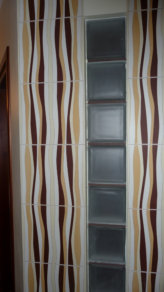 Ściana wyłożona płytkami z falistym wzorem w odcieniach brązu, beżu i bieli, sąsiadująca z pionowym pasem z luksferów w łazience.