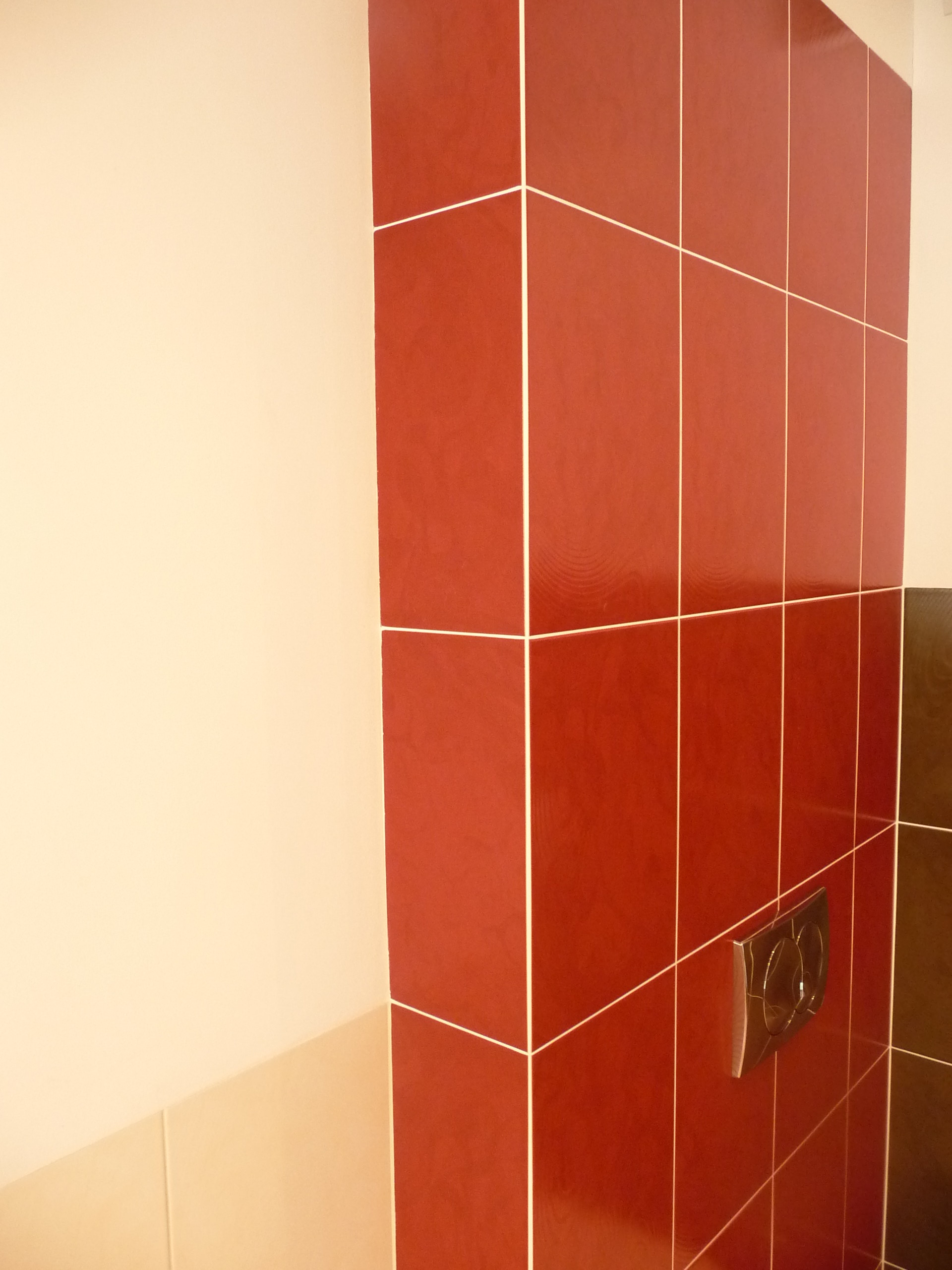 Czerwone, błyszczące płytki ceramiczne ułożone na ścianie łazienki z widocznym przyciskiem spłuczki i fragmentem sąsiedniej ściany pomalowanej na beżowo.
