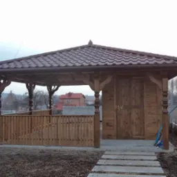 Drewniana altana z brązowym dachem z dachówki, zamykana na drewniane drzwi, z ozdobnymi kolumnami i niskim drewnianym płotkiem, na nierównym terenie z betonowymi płytami chodnikowymi.