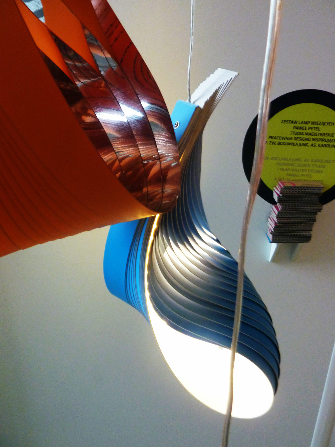 Wielobarwna lampa wisząca z pomarańczowymi i niebieskimi elementami, wykonana z warstwowych materiałów, oświetlona od wewnątrz, z widocznym plakatem informacyjnym w tle.