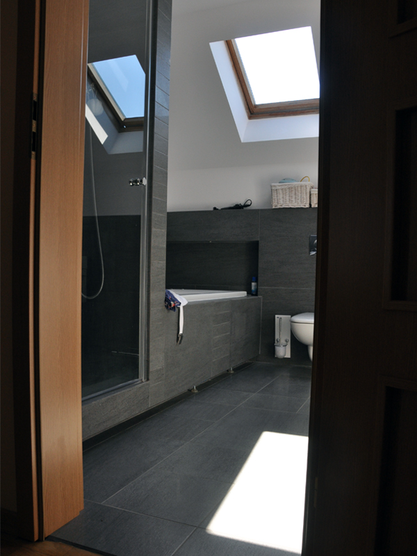 Nowoczesna łazienka z szarą glazurą, kabiną prysznicową i wanną pod skosem dachu, widok zza otwartych drewnianych drzwi, wpadające promienie słoneczne na podłodze.