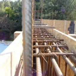 Zbrojenie w szalunku z desek, widok z góry na konstrukcję z prętów stalowych połączonych drutem, w tle fragment basenu i zieleń ogrodu.