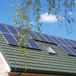 Dom z zielonym dachem pokrytym panelami fotowoltaicznymi i oknem dachowym, na tle błękitnego nieba z delikatnymi chmurami i gałęzi drzewa na pierwszym planie.