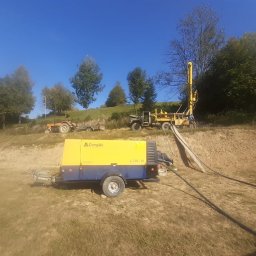 WITOLD STOPYRA FHU. WIT-WIERT - Żółta wiertnica na pagórkowatym terenie, z widocznym traktorem w tle, podczas wiercenia studni w słoneczny dzień.