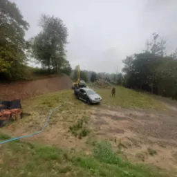 Wiercenie studni głębinowej na nierównym terenie: wiertnica na podwoziu samochodu ciężarowego, stos gruzu, widoczny pracownik w oddali, błękitny wąż doprowadzający wodę.