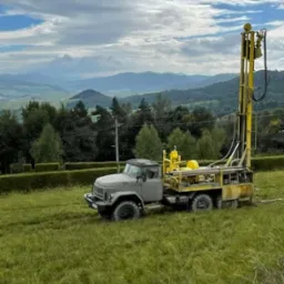 Ciężarówka wiertnicza z żółtym masztem na zielonym polu z widokiem na zalesione wzgórza i zachmurzone niebo.