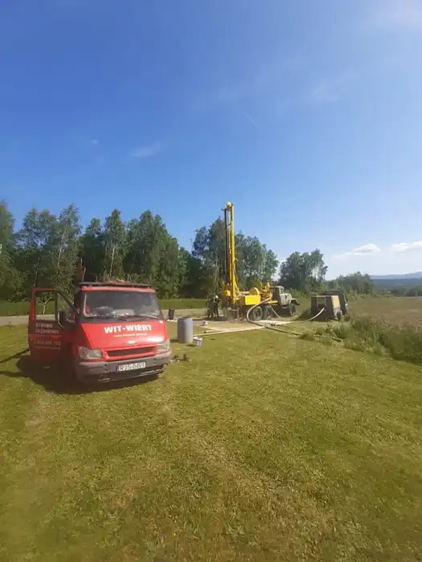 Czerwony samochód dostawczy firmy WIT-WIERT z otwartymi drzwiami na trawniku, w tle żółta wiertnica podczas pracy przy wierceniu studni w słoneczny dzień.