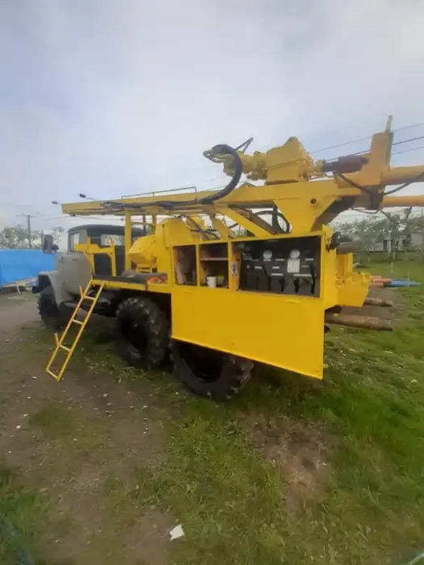 Żółta ciężarówka wiertnicza z drabinką, stojąca na trawiastym terenie, ze sprzętem wiertniczym na górze i rurami wiertniczymi obok.