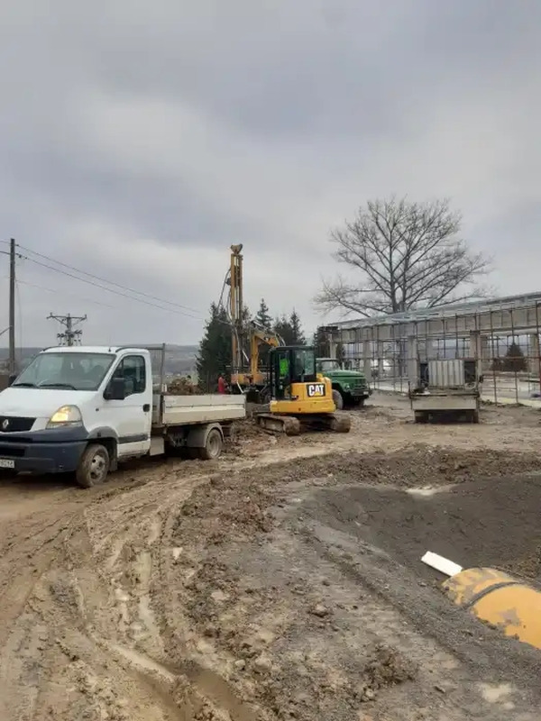 Prace ziemne z użyciem wiertnicy i koparki CAT na placu budowy w pochmurny dzień, widoczny biały samochód dostawczy i częściowo zarys budynku w tle.