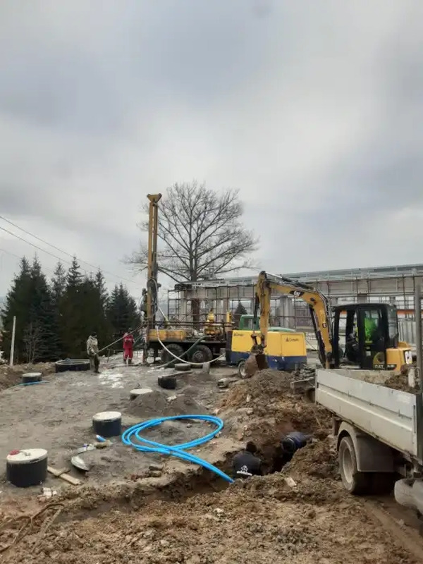 Wiercenie studni: żółta wiertnica z masztem na placu budowy, obok wykopu z niebieskimi rurami, widoczny koparka i ciężarówka, pochmurne niebo.