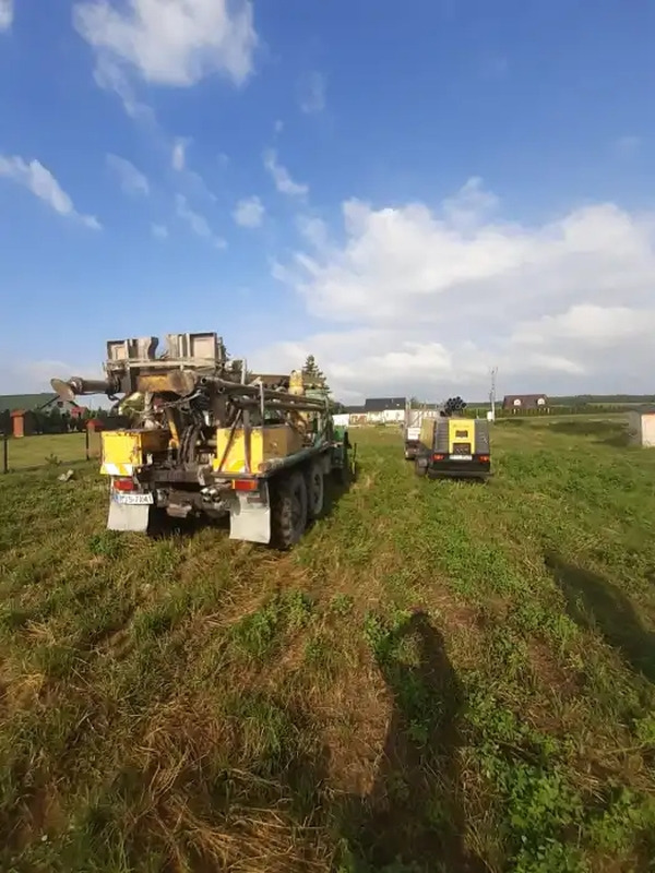 Żółty samochód ciężarowy z wiertnicą na łące, mniejszy pojazd w tle, błękitne niebo z chmurami.