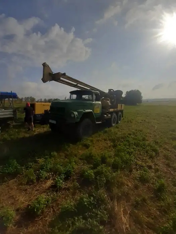 Zielony samochód ciężarowy z wiertnicą na łące, prawdopodobnie podczas wiercenia studni; widoczne niebo z chmurami i silne słońce w prawym górnym rogu.