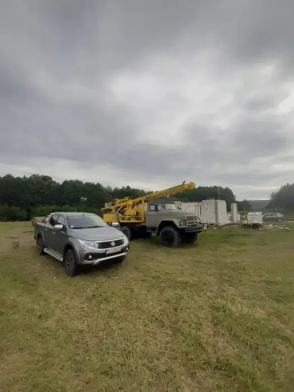 Szara ciężarówka z żółtym masztem wiertniczym na trawiastym terenie budowy, obok zaparkowany szary pickup, widoczne bloczki fundamentowe w tle.