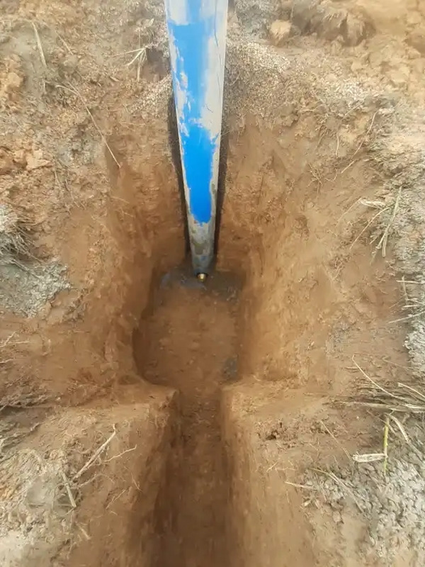 Niebieska rura studni głębinowej wystająca z wykopu w ziemi, widoczny mosiężny zawór na dole rury.