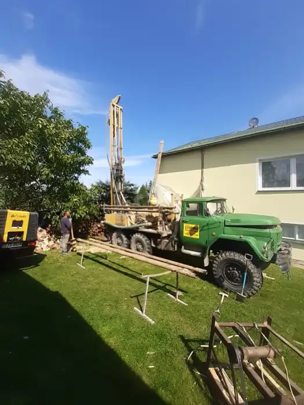 Wiertnica zamontowana na zielonej ciężarówce podczas pracy na trawniku obok budynku, widoczne rury wiertnicze na stojakach i pracownik w tle.