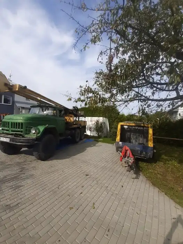 Zielony samochód ciężarowy z wiertnicą na podwórku z kostki brukowej, obok zaparkowana żółto-niebieska przyczepa, w tle dom i drzewa.