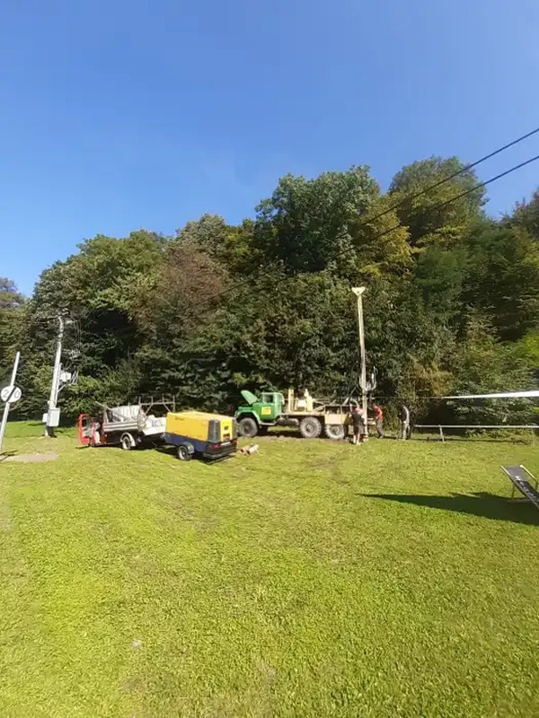 Wiercenie otworu w ziemi przy użyciu ciężkiego sprzętu na zewnątrz, widoczne drzewa i niebieskie niebo.