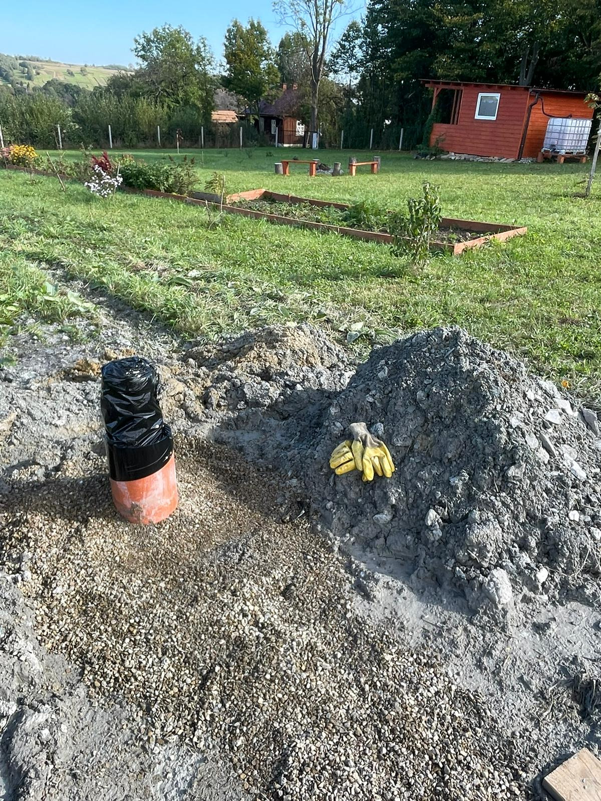 Pomarańczowa rura z czarnym zabezpieczeniem wystaje z ziemi, obok leżą żółte rękawice robocze na stercie ziemi i żwiru, w tle ogród z domkami.