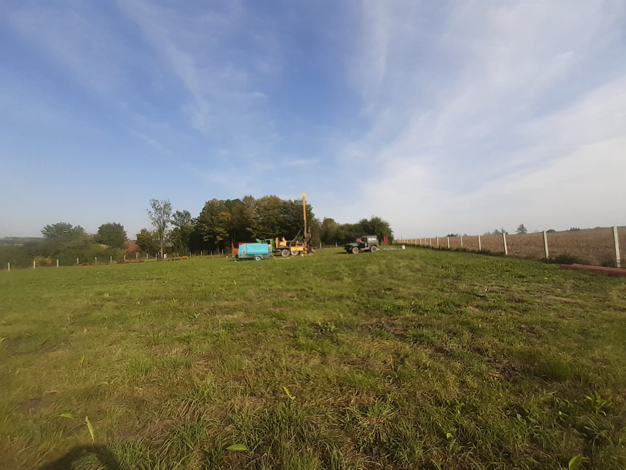 Sprzęt do wiercenia na zewnątrz, na zielonym terenie, widoczny wiertnica, cysterna z wodą i samochód terenowy, całość otoczona ogrodzeniem z betonowych słupków.