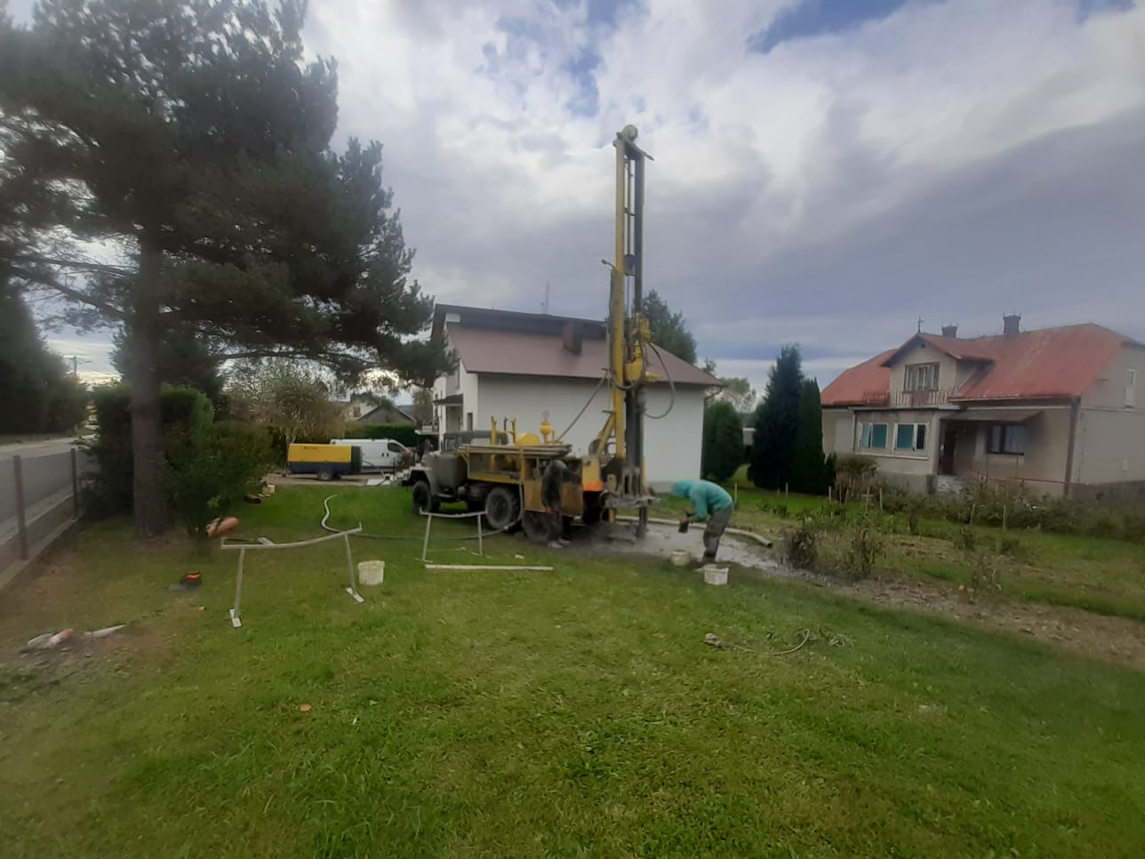 Wiercenie studni głębinowej w Gliniczku przy użyciu ciężkiego sprzętu wiertniczego na tle domów jednorodzinnych. Widoczny pracownik w kombinezonie roboczym i pomocniczy agregat prądotwórczy.