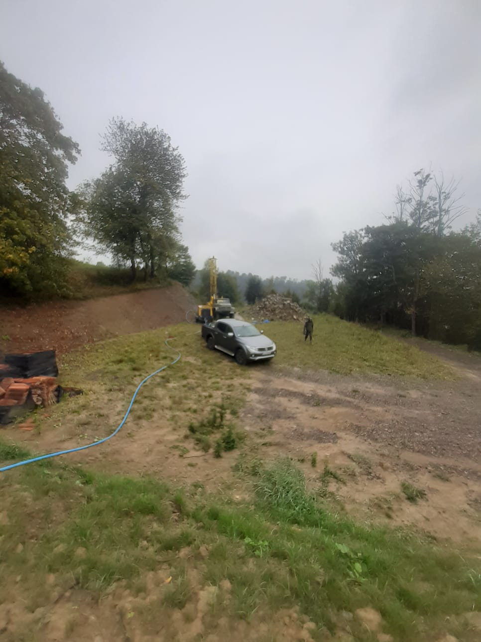 Wiercenie studni głębinowej na nierównym terenie: wiertnica na podwoziu samochodu ciężarowego, stos gruzu, widoczny pracownik w oddali, błękitny wąż doprowadzający wodę.