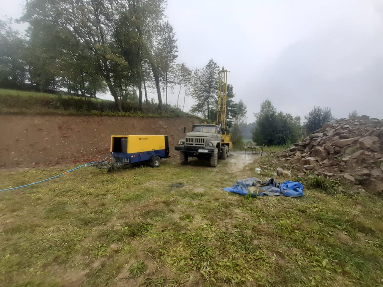 Prace wiertnicze w terenie: duża wiertnica samochodowa ustawiona na trawiastym terenie, obok agregat prądotwórczy, w tle widoczny wykop i sterta kamieni.