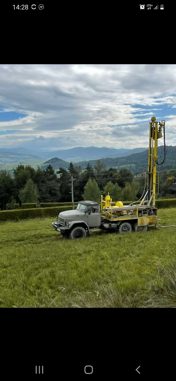 Ciężarówka wiertnicza z żółtym masztem na zielonym polu z widokiem na zalesione wzgórza i zachmurzone niebo.