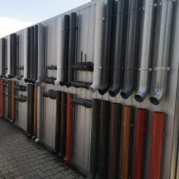 Ekspozycja różnokolorowych rynien spustowych z PCV i metalu, zamocowanych na szarej, blaszanej ścianie, prezentacja w składzie budowlanym.