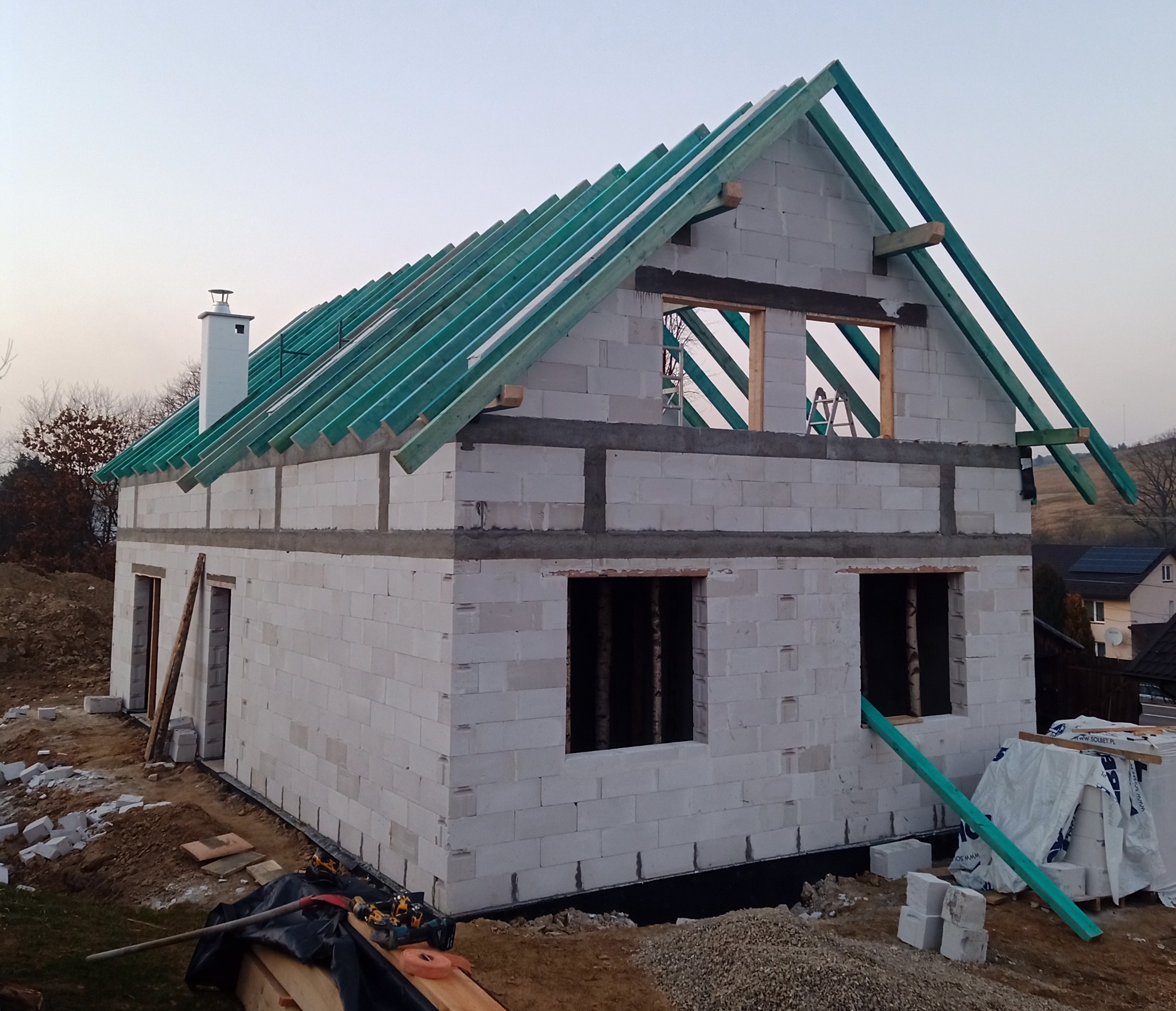 Surowy stan domu z białych bloczków, widoczne zielone elementy konstrukcji dachu, komin i otwory okienne bez okien, teren budowy z narzędziami i materiałami.