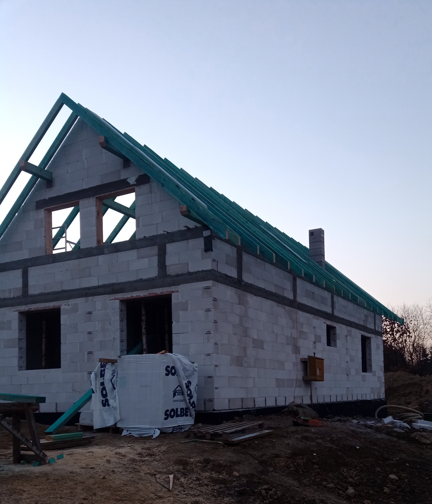 Wznoszenie surowego domu murowanego z bloczków w Birczy, widoczne elementy konstrukcji dachu i komina, materiały budowlane składowane obok.