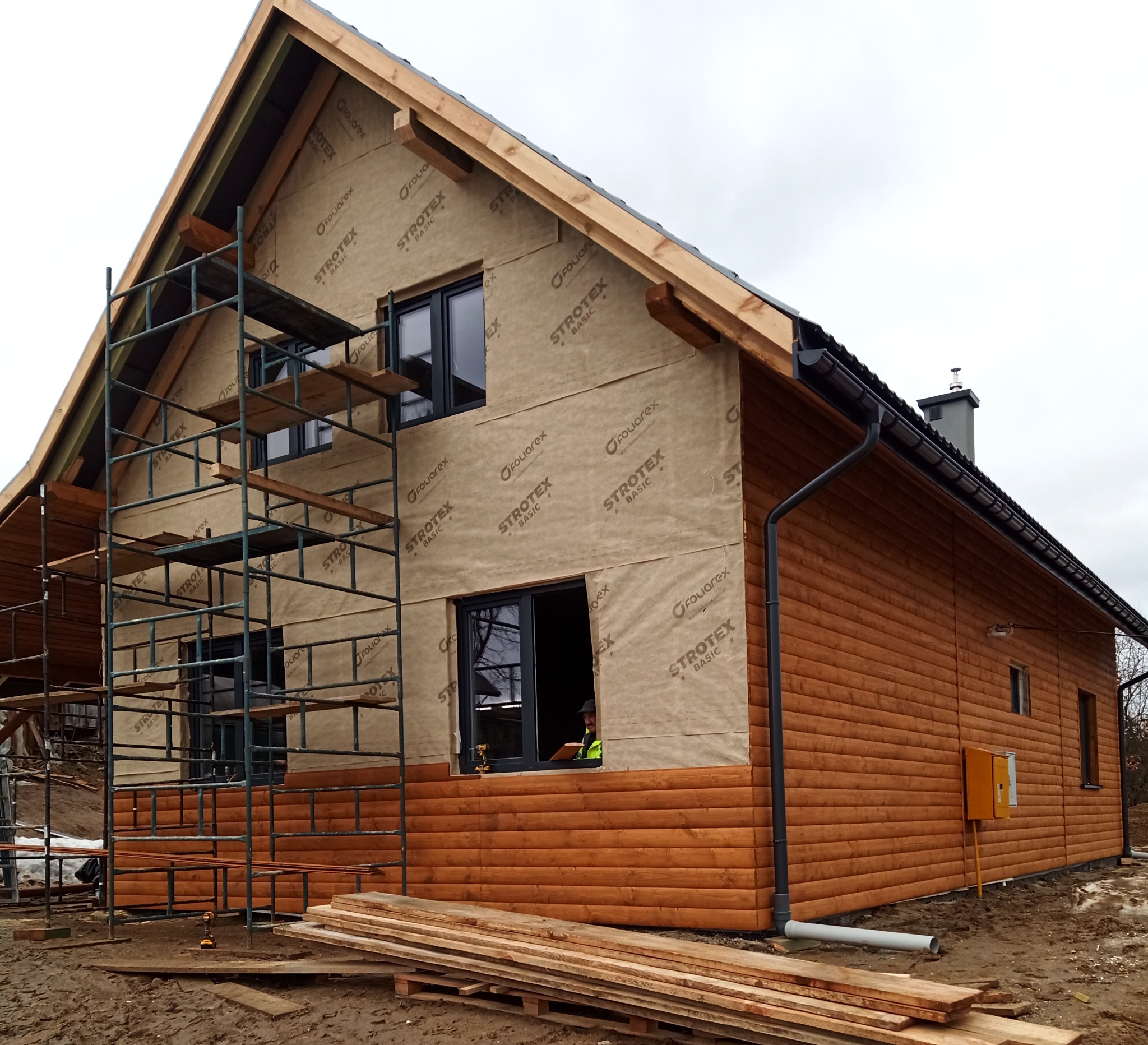 Dom w trakcie budowy, z widocznym ociepleniem ścian zewnętrznych folią ochronną, rusztowaniem i częściowo wykończoną elewacją drewnianą, widoczny pracownik w oknie.