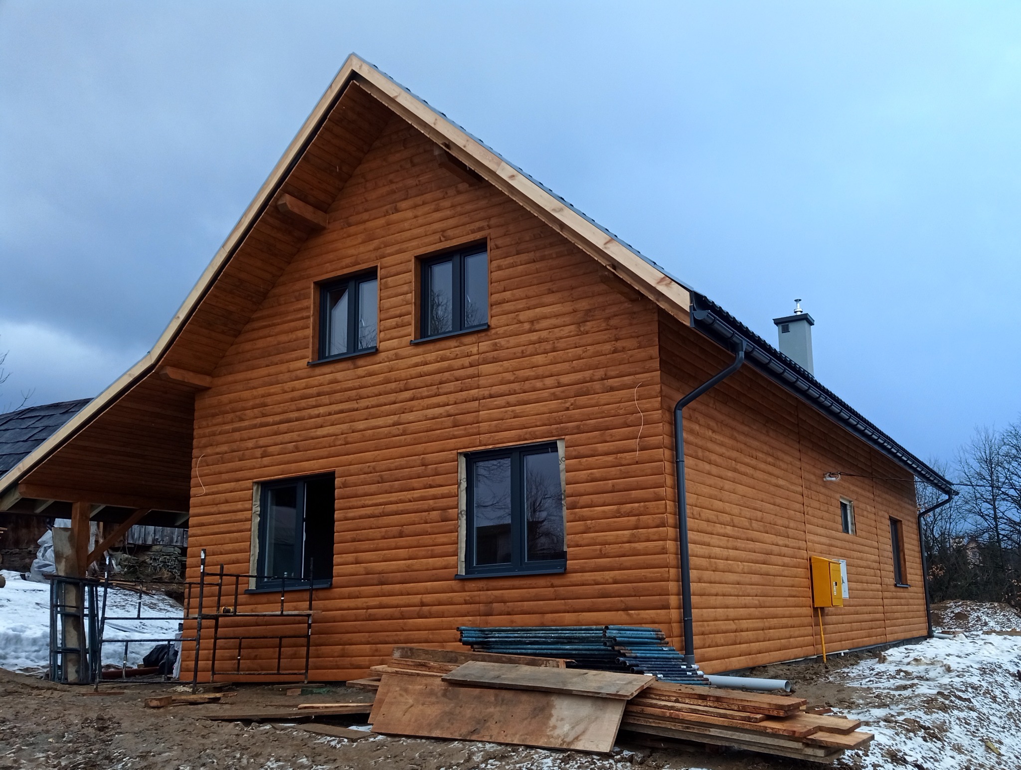 Nowo wybudowany, dwukondygnacyjny dom z drewnianą elewacją w trakcie wykańczania, widoczne rusztowania, składowane deski oraz skrzynka gazowa na elewacji bocznej