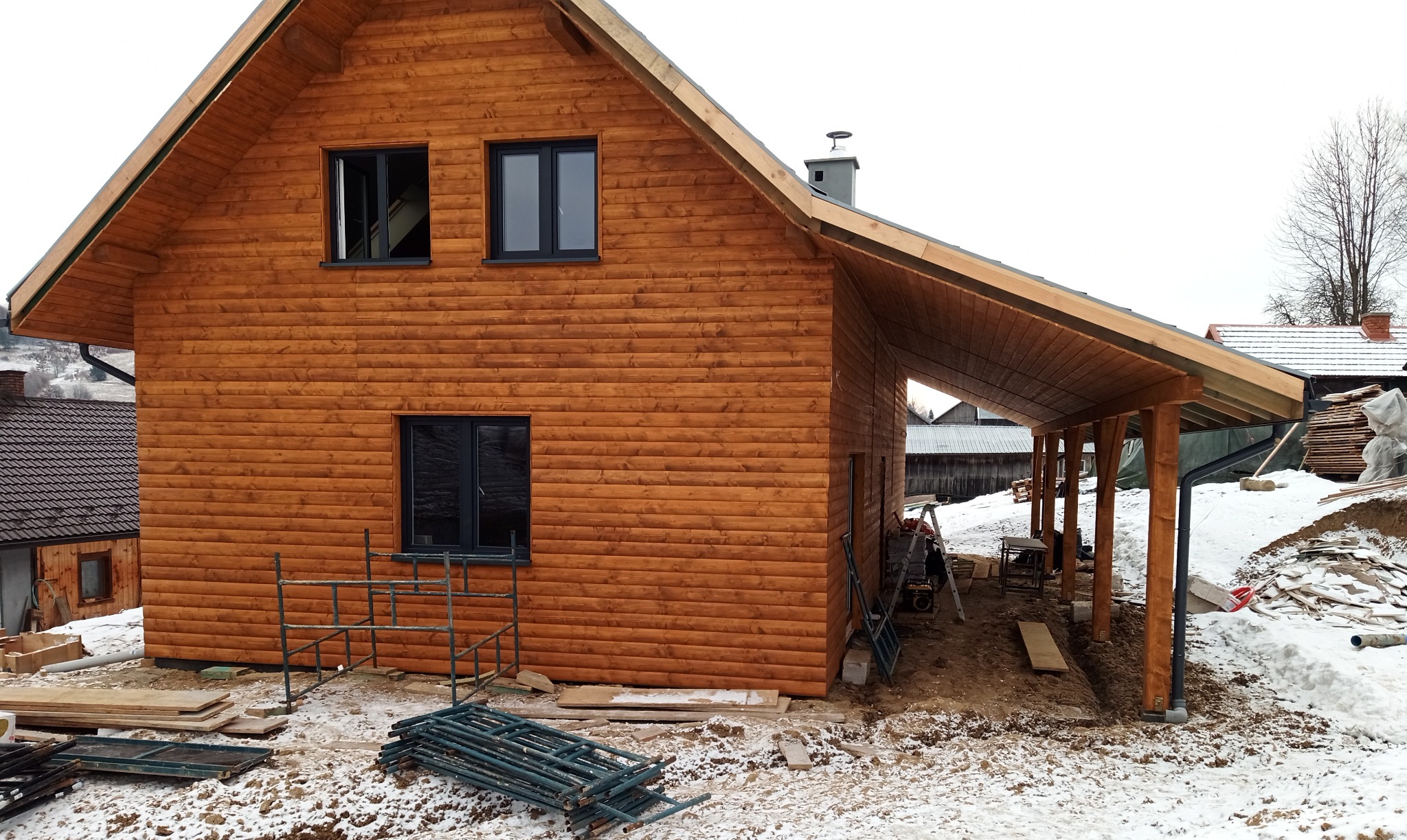 Nowo wybudowany, dwukondygnacyjny dom z drewna, z dobudowanym zadaszeniem, otoczony śniegiem i materiałami budowlanymi, widoczne rusztowania.